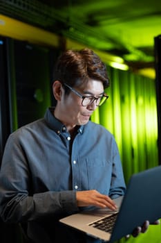 Asian male engineer using laptop in computer server room. database server management and maintenance concept