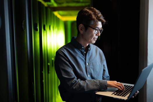 Asian male engineer using laptop in computer server room. database server management and maintenance concept