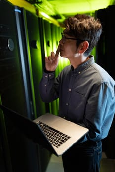 Thoughtful asian male engineer with laptop inspecting in computer server room. database server management and maintenance concept