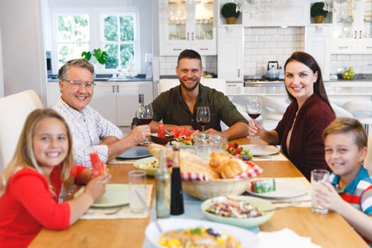 Portrait of caucasian grandfather and parents with son and daughter sitting at table having dinner. family spending time together at home.