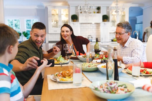 Caucasian grandfather and parents with daughter and son showing phone while having dinner. family spending time together at home.