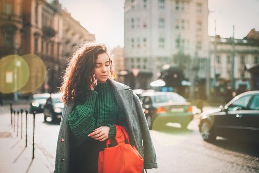 A woman on the street uses a mobile phone. online shopping. use of mobile applications. beautiful young woman with long curly dark hair in a casual coat, trendy green sweater and red handbag