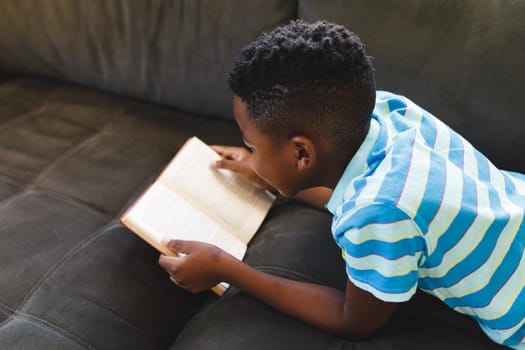 African american boy reading book and lying on couch in living room. spending time alone at home.