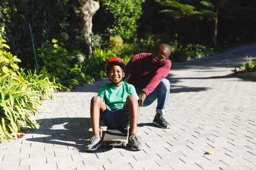 Portrait of african american father with son smiling and playing with skateboard in garden. family spending time at home.