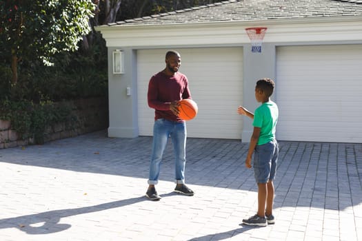 Smiling african american father and son playing basketball in sunny garden. family spending time at home.