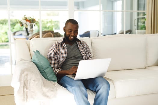 Smiling african american man using laptop and sitting on couch in living room. spending time alone at home with technology.