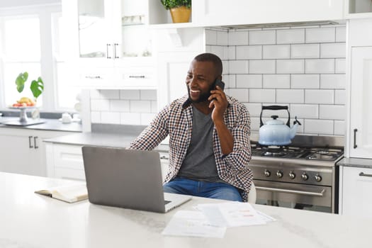 Smiling african american man sitting in kitchen working using laptop and smartphone. remote working from home with technology.