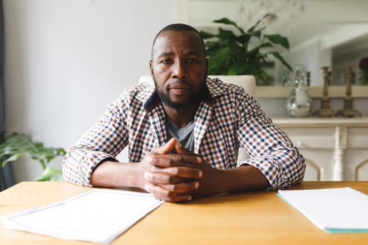 Serious african american man sitting at table in dining room listening during video call. flexible working from home with technology.