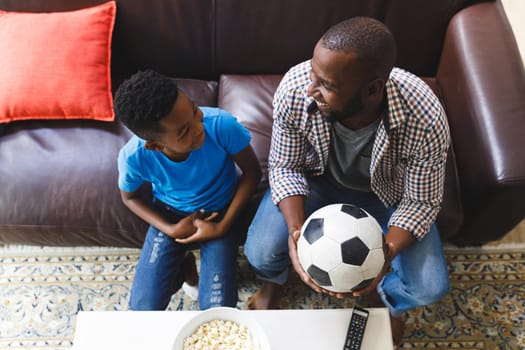 Smiling african american father and son sitting on sofa, watching match on tv and talking. family spending time together at home.