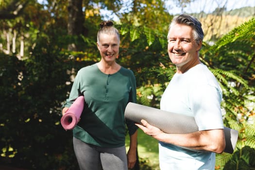 Portrait of happy senior caucasian couple holding yoga mats, looking to camera in sunny garden. healthy retirement lifestyle, spending time at home.