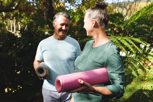 Happy senior caucasian couple practicing yoga, holding yoga mats in sunny garden. healthy retirement lifestyle, spending time at home.