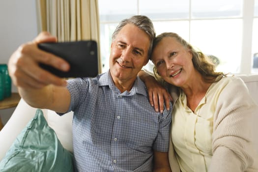 Happy senior caucasian couple in living room, sitting on sofa, taking selfies and smiling. retirement lifestyle, spending time at home.