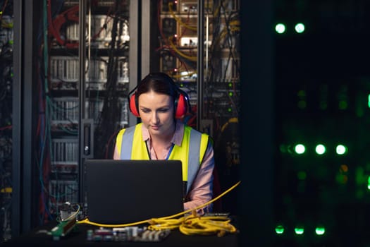 Caucasian female engineer wearing ear plugs using a laptop in computer server room. database server management and maintenance concept