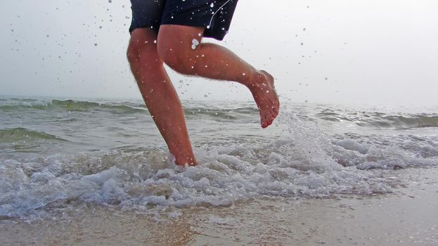 The feet of a man running along a sea beach in foggy weather. Splashes from the waves are flying around. sand and waves under the feet of a man running in shorts. Barefoot morning jogging on the beach