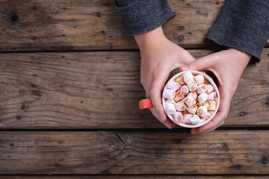 Female hands holding mug of hot chocolate drink filled with marshmallows at table with copy space. lifestyle and cafe culture.