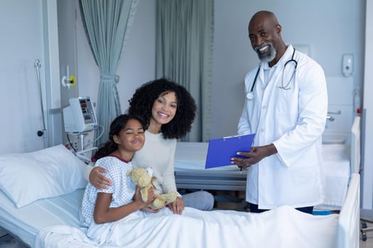 Smiling african american male doctor with mixed race mother and her sick daughter, in hospital bed. medicine, health and healthcare services.