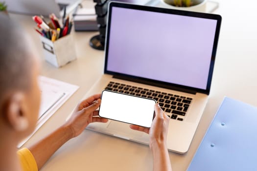 African american businesswoman in casual using smartphone in front of laptop with copy space. business, wireless technology and office.