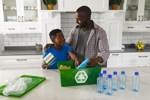 Happy african american father and son in kitchen talking and sorting rubbish for recycling. family spending time at home.