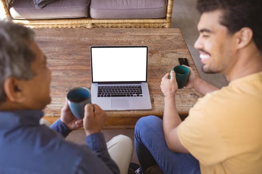 Happy biracial adult son and senior father holding cups of coffe and making laptop video call. family time at home using technology together.