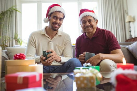 Portrait of happy biracial adult son and senior father in santa hats making christmas video call. christmas, festivity and communication technology.