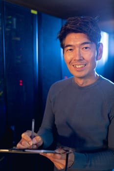 Portrait of asian male engineer smiling while writing on clipboard in computer server room. database server management and maintenance concept