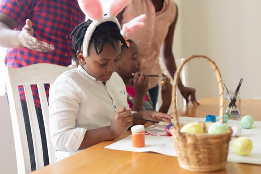 Happy african american parents with son and daughter in bunny ears painting colourful eggs. celebrating easter at home in isolation during quarantine lockdown.