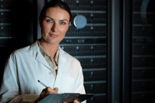 Portrait of caucasian female engineer wearing an apron writing on clipboard in computer server room. database server management and maintenance concept