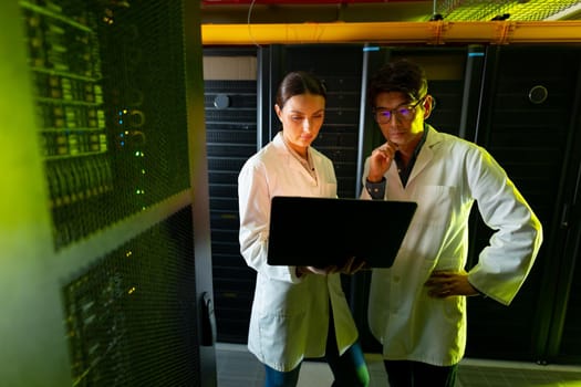 Diverse male and female engineers wearing aprons using laptop while inspecting in server room. database server management and maintenance concept