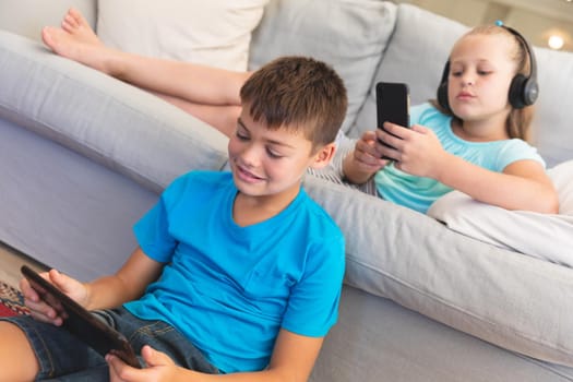 Caucasian brother and sister wearing headphones and using tablets at home. childhood with technology, spending free time at home.