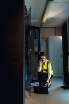 Caucasian female engineer with laptop talking on smartphone while inspecting in computer server room. database server management and maintenance concept