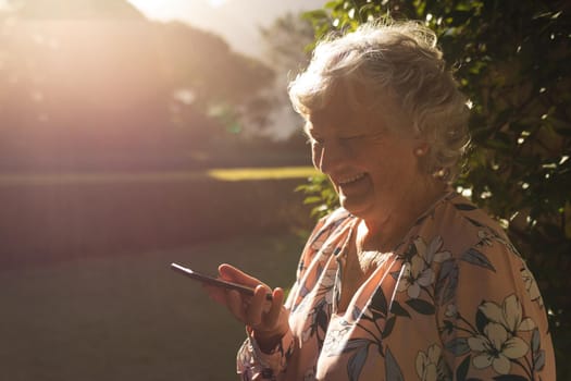 Smiling senior caucasian woman using smartphone in sunny garden. luxury retirement lifestyle, spending time alone at home.