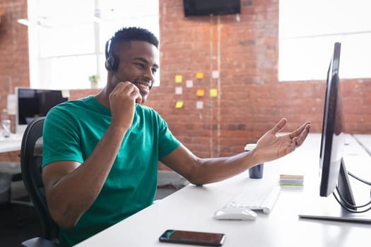 African american businessman having video call sitting in front of computer using headphones. independent creative design business.