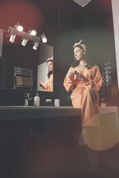 Portrait view of beautiful young woman getting ready in home bathroom decorated with black tiles. moisturising cream. female cosmetics, mirror face reflection.