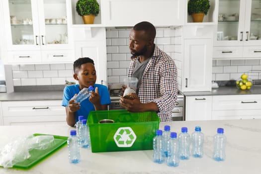 Happy african american father and son in kitchen talking and sorting plastic waste for recycling. family spending time at home.