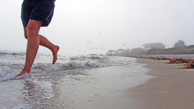 Splashes from the waves are flying around. sand and waves under the feet of a man running in shorts. Barefoot morning jogging on the beach. The feet of a man running along a sea beach in foggy weather.