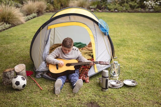 Caucasian boy playing guitar while sitting in a tent in the garden. childhood and hobby concept