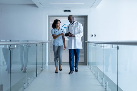 Diverse male and female doctor walking in hospital corridor talking and looking at tablet. medicine, health and healthcare services.