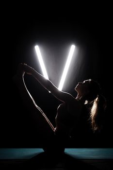 Fit woman practicing yoga poses. Silhouette girl doing exercise in studio against black background with v shaped white led tube light. No stress inner balance concept.