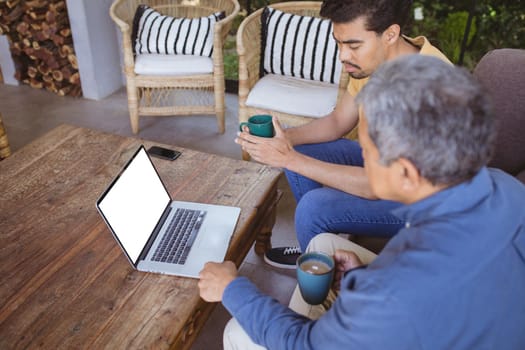 Biracial adult son and senior father holding cups of coffe and making laptop video call. family time at home using technology together.