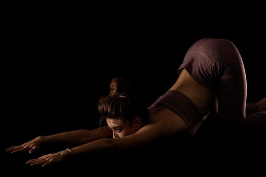 Fit woman practicing yoga poses. Side lit half silhouette girl doing exercise in studio against black background.