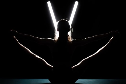 Fit woman practicing yoga poses. Silhouette girl doing exercise in studio against black background with v shaped white led tube light. No stress inner balance concept.