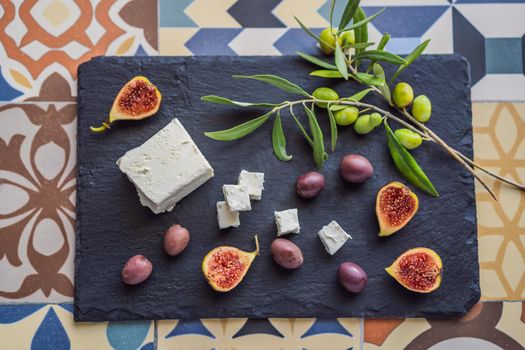 Green and black olives with loaf of fresh bread, feta cheese and young olives branch on olive wood chopping board over dark background.