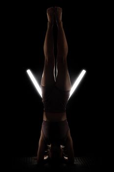 Fit woman practicing yoga poses. Silhouette girl doing exercise in studio against black background with v shaped white led tube light. No stress inner balance concept.