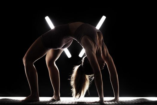 Fit woman practicing yoga poses. Silhouette girl doing exercise in studio against black background with v shaped white led tube light. No stress inner balance concept.