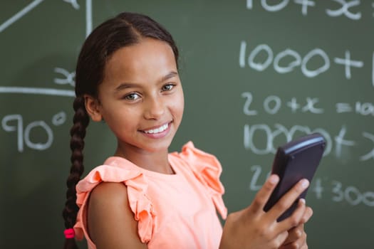 Portrait of smiling mixed race schoolgirl standing at chalkboard in classroom using smartphone. childhood, technology and education at elementary school.