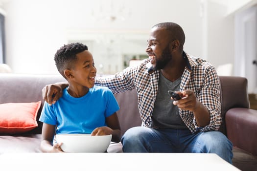 Happy african american father and son sitting on sofa, watching tv. family spending time together at home.