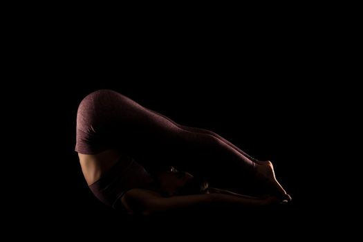 Fit woman practicing yoga poses. Side lit half silhouette girl doing exercise in studio against black background.