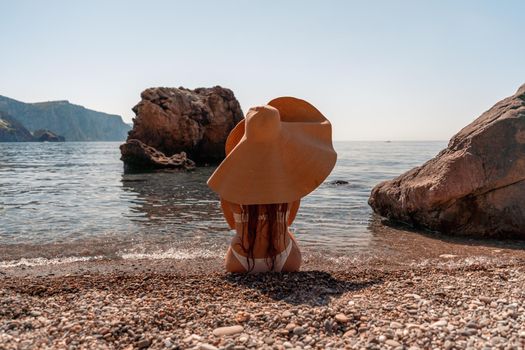 Tanned middle-aged woman with long hair and a white bathing suit. He sits on the seashore in a large sun hat with his back and looks at the sea