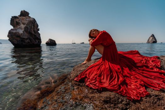 Beautiful sensual woman in a flying red dress and long hair, sitting on a rock above the beautiful sea in a large bay