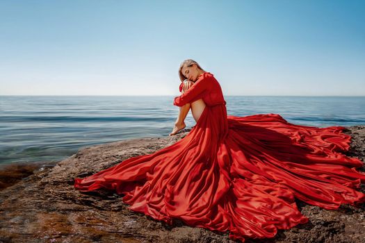 Beautiful sensual woman in a flying red dress and long hair, sitting on a rock above the beautiful sea in a large bay